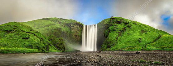 Wzornik fototapety z widokiem na wodospad wypływający z zielonych gór na Islandii. Fototapeta do pokoju dziennego, sypialni, salonu, biura, gabinetu, przedpokoju i jadalni.