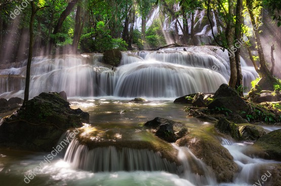 Wzornik fototapety z pięknym wodospadem Pha Tad w Parku Narodowym w Tajlandii. Promienie słoneczne przebijają się przez gęstwinę zielonych drzew. Fototapeta przeznaczona do salony, sypialni, pokoju młodzieżowego, gabinetu czy biura.