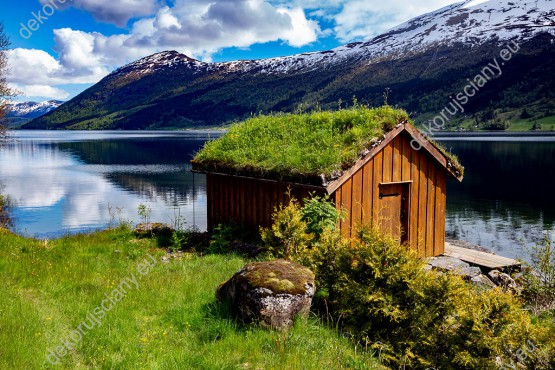 Wzornik fototapety z widokiem na drewnianą chatkę przy jeziorze z trawiastym dywanem na dachu, na tle gór Norwegii. Fototapeta do pokoju dziennego, sypialni, salonu, biura, gabinetu, przedpokoju i jadalni.