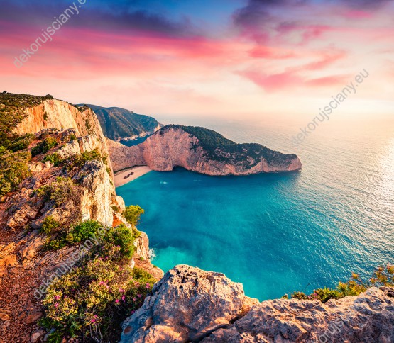 Wzornik fototapety z widokiem na piękny krajobraz skalnego wybrzeża i lazurowej wody przy plaży Shipwreck, w Grecji w kolorach zachodzącego słońca. Fototapeta do pokoju dziennego, sypialni, salonu, biura, gabinetu, przedpokoju i jadalni.