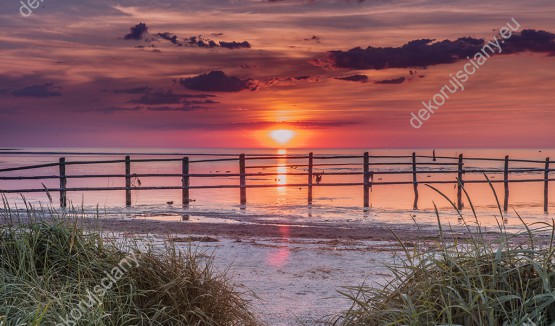 Wzornik fototapety widokiem na piaszczystą plażę i morze zabarwione kolorami zachodzącego słońca. Fototapeta do pokoju dziennego, sypialni, salonu, biura, gabinetu, przedpokoju i jadalni.