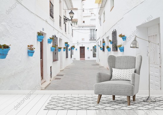 Wizualizacja fototapety z widokiem ulicy miasta w Andaluzji, w Hiszpanii. Białe domy zdobią niebieskie doniczki z kwiatami. Fototapeta przeznaczona do salonu, sypialni, pokoju dziennego, gabinetu, biura, przedpokoju, optycznie powiększa pomieszczenie.