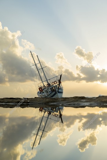 Wzornik fototapety z widokiem na żaglówkę na brzegu kamienistej plaży, odbijającej się lustrze wody. Fototapeta do pokoju dziennego, salonu, biura, gabinetu, sypialni, przedpokoju i jadalni.