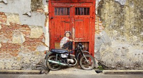 Wzornik fototapety z kategorii murali przedstawia motocyklistę siedzącego na motorze, na tle starych, czerwonych drzwi. Fototapeta do pokoju dziennego, młodzieżowego, sypialni, salonu, biura, gabinetu, przedpokoju i jadalni.